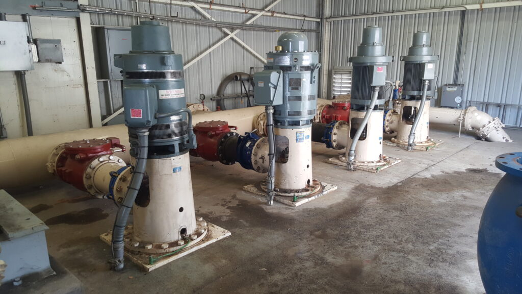 Four vertical pumps with motors connected to large pipes in a pump house