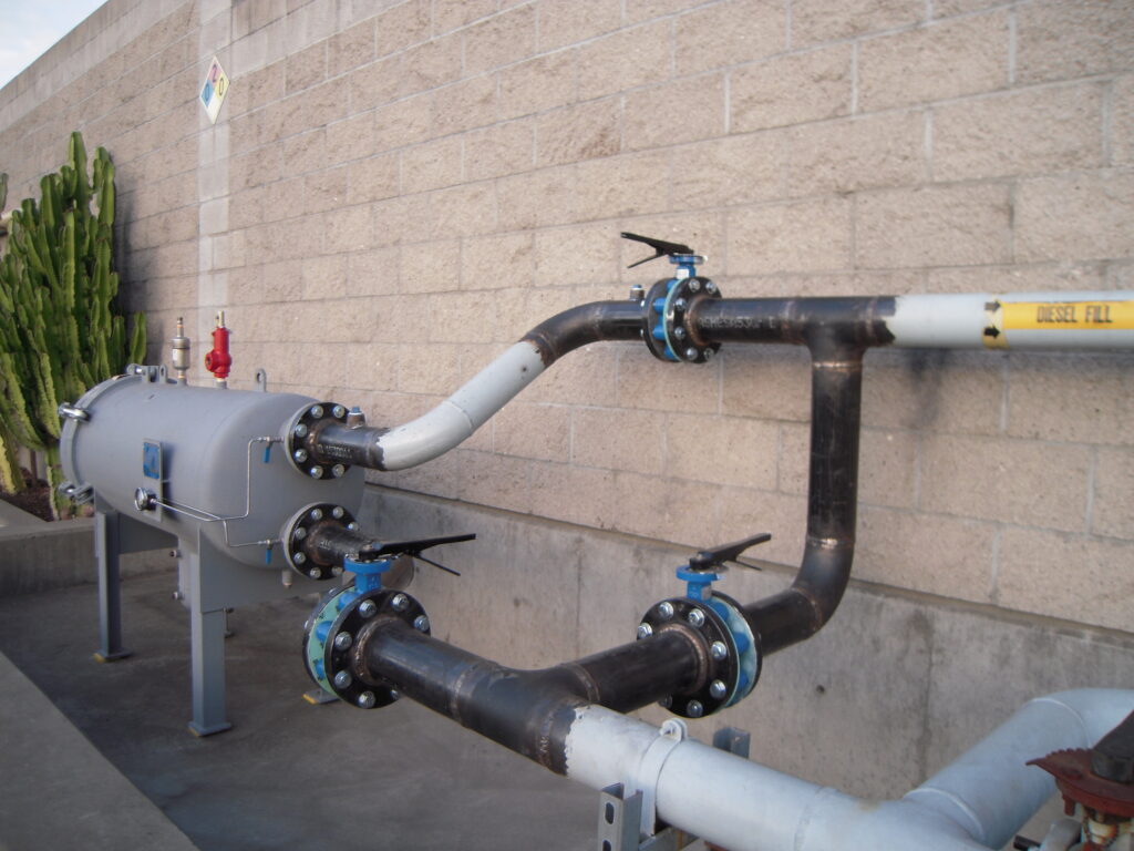 Black steel diesel fill piping with blue valves and a filter housing along a block wall