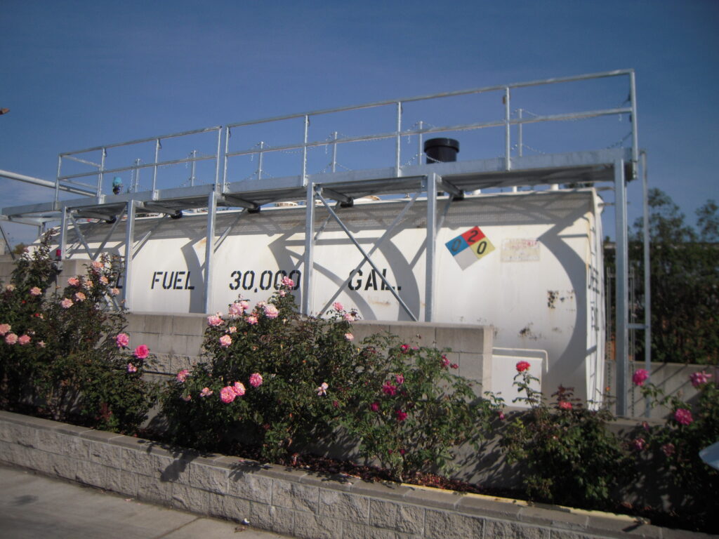 White 30,000-gallon fuel tank with a steel access platform on top, behind rose bushes